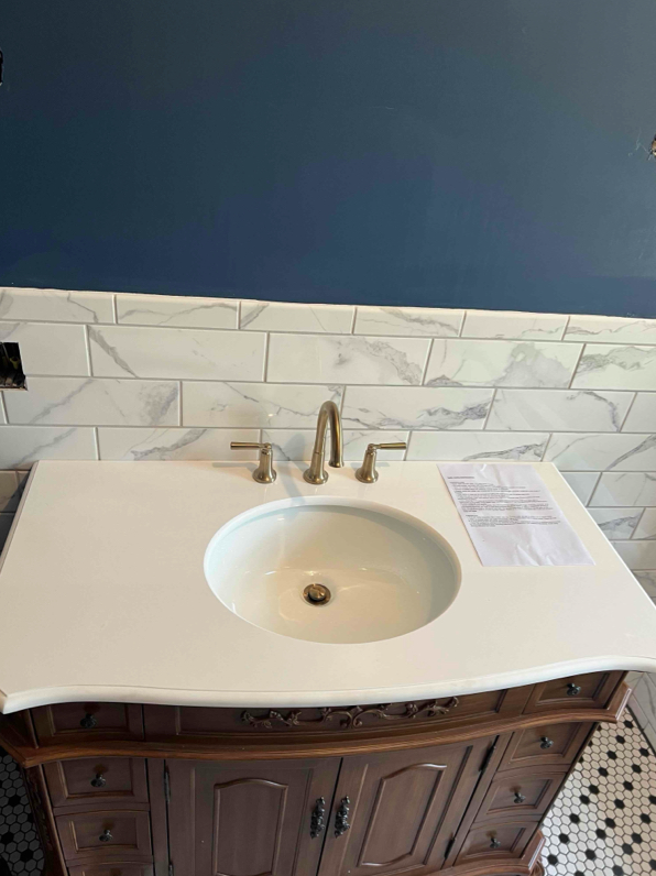 Bathroom vanity with white countertop, round sink, and gold faucet, set against blue wall and tile backsplash.
