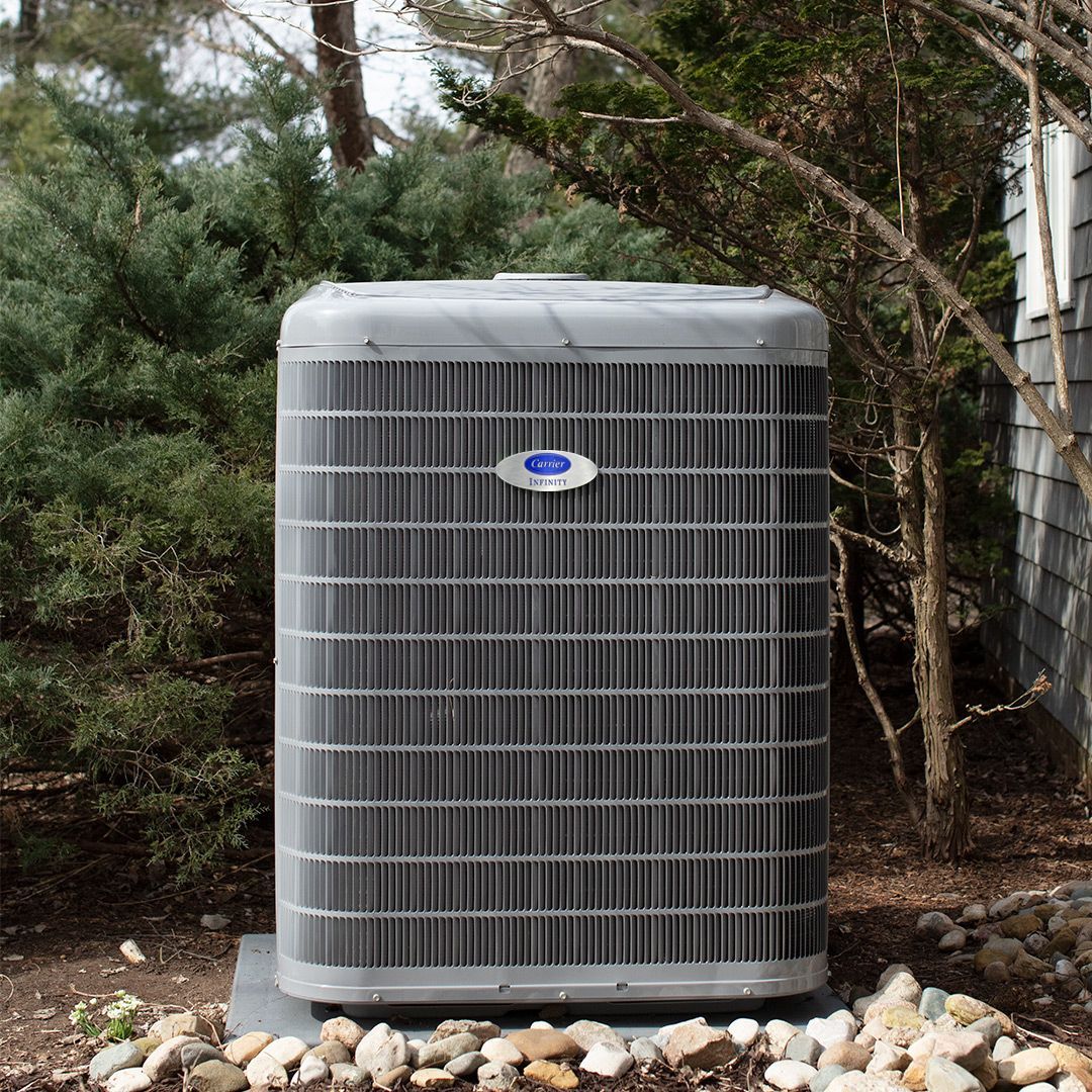 Outdoor air conditioning unit with gray metal casing, surrounded by shrubs and small rocks.
