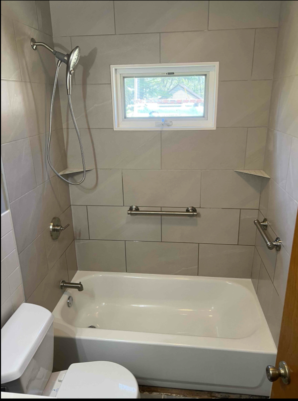 Bathroom with white tub, toilet, light gray tile, grab bars, and window.