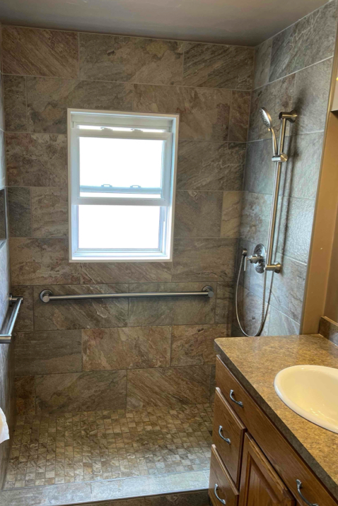 Bathroom with tiled shower and grab bar, window, and vanity.