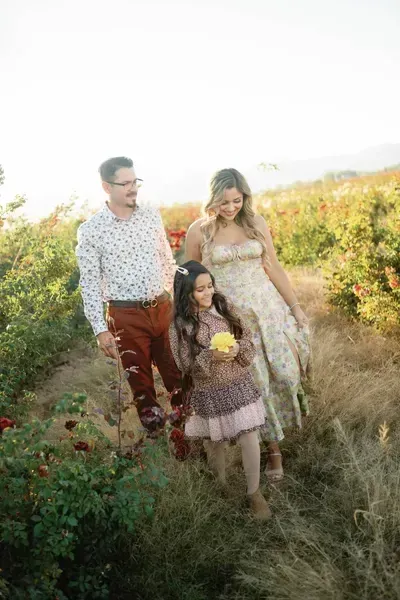 Three people walking through a grassy field in bright sunlight, one holding a yellow flower