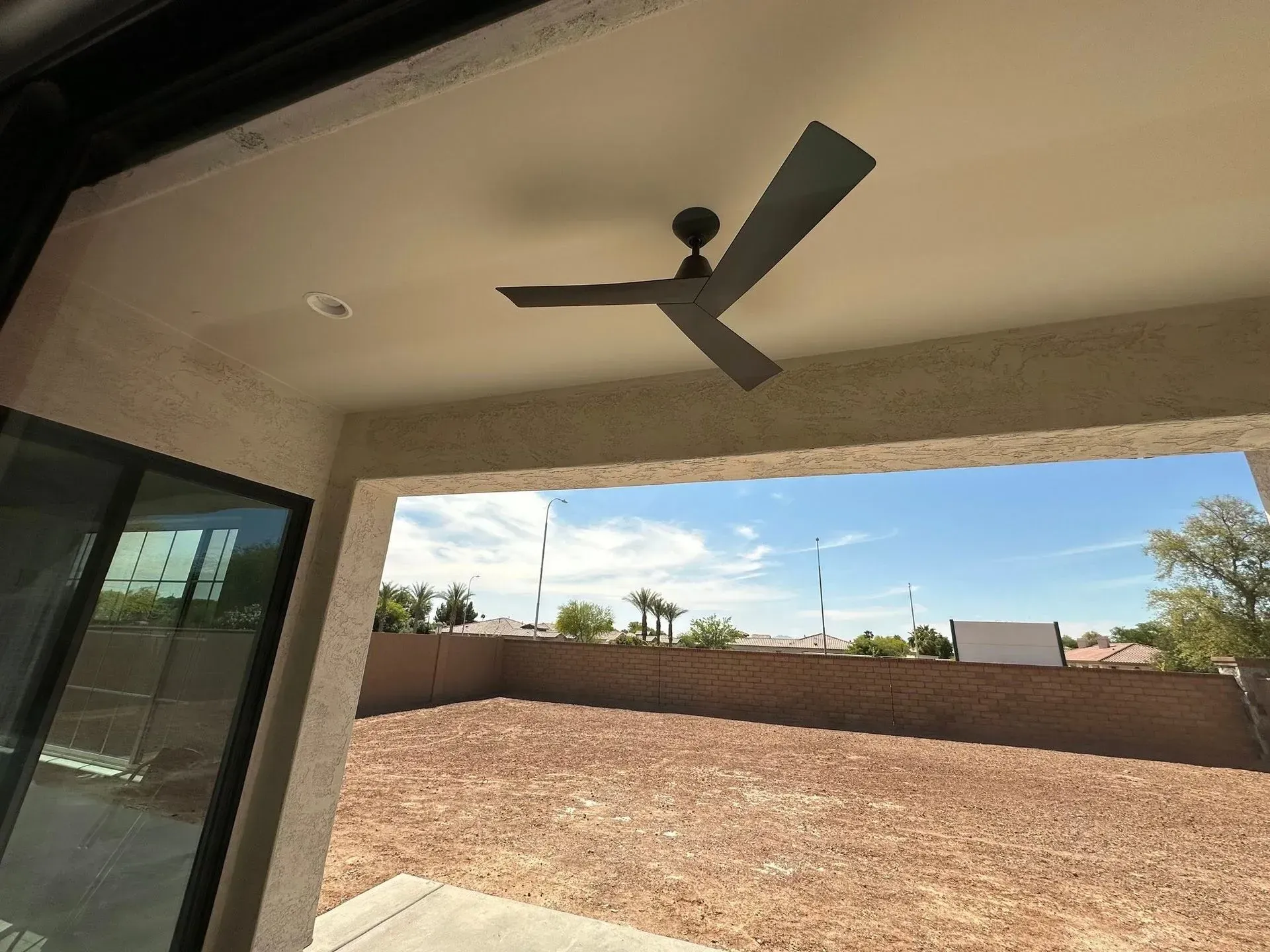 Covered patio with a ceiling fan, sliding glass door, and a fenced dirt backyard under a blue sky.