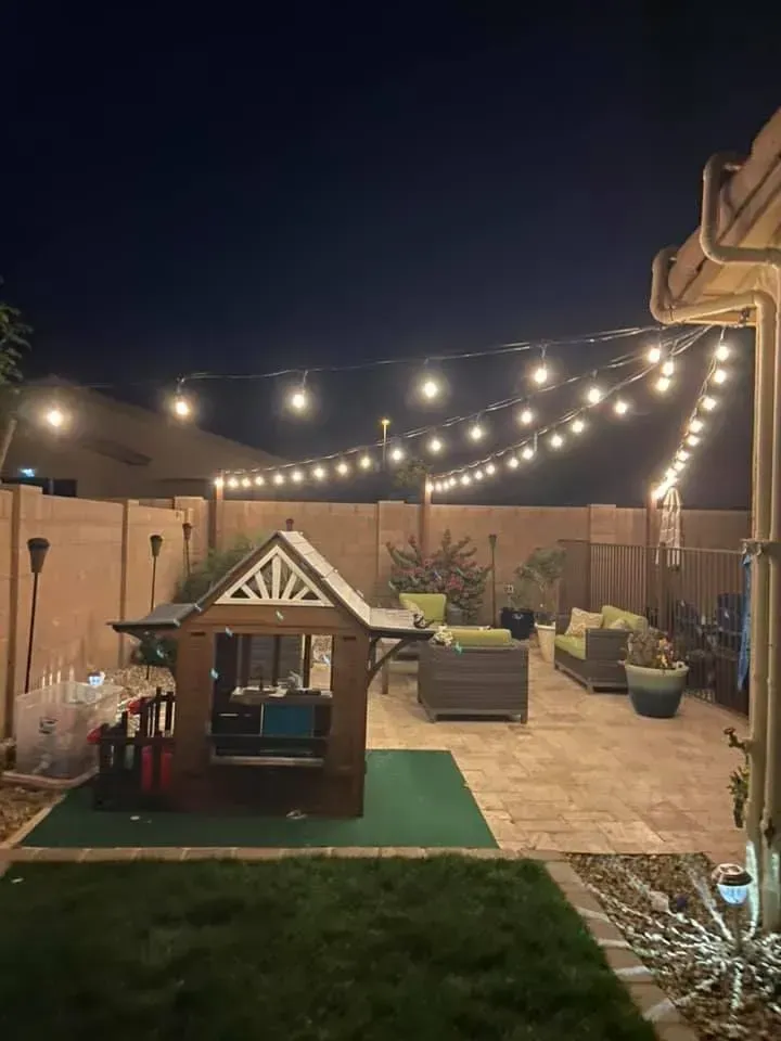 Backyard patio at night with string lights, seating area, and a small playhouse on a green mat