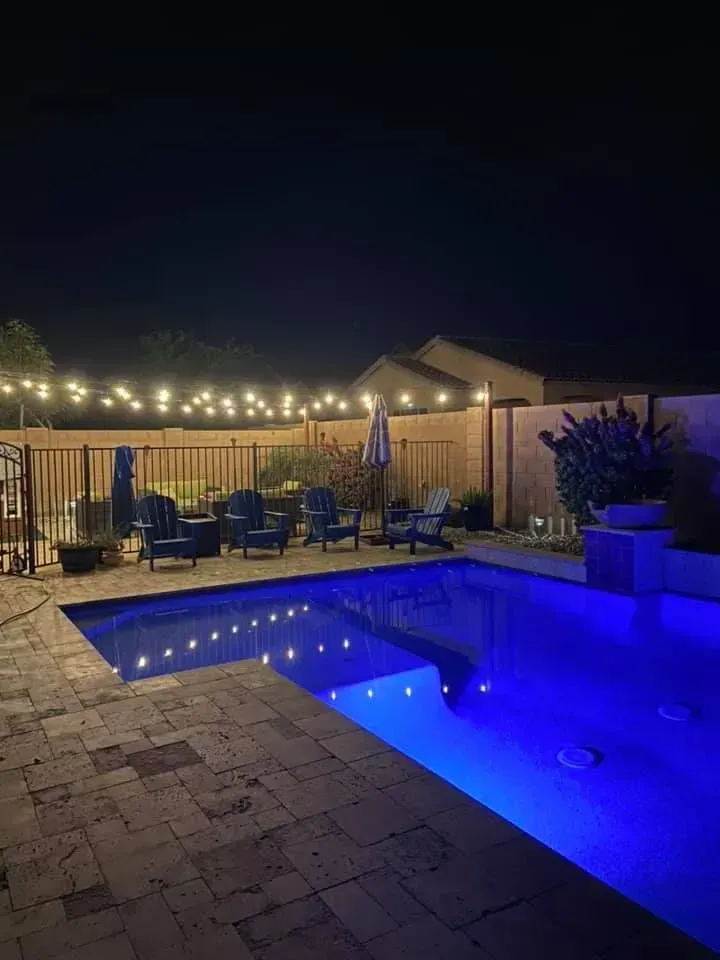 Nighttime backyard with lit swimming pool, patio seating, and string lights under a dark sky