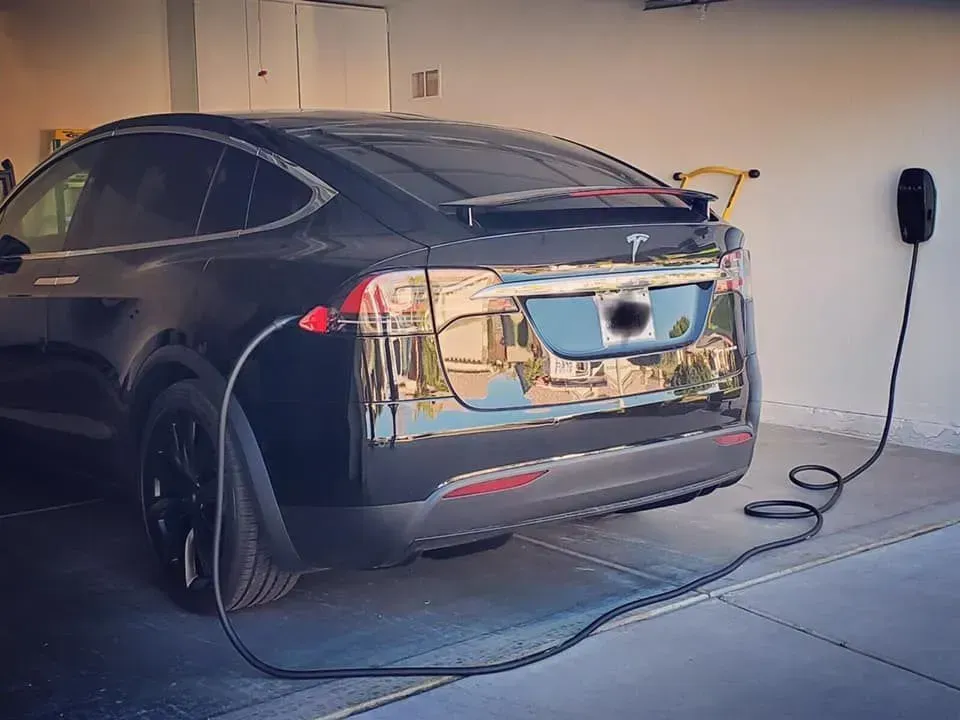 Black Tesla plugged into a wall charger in a garage, with an extension cord on the floor