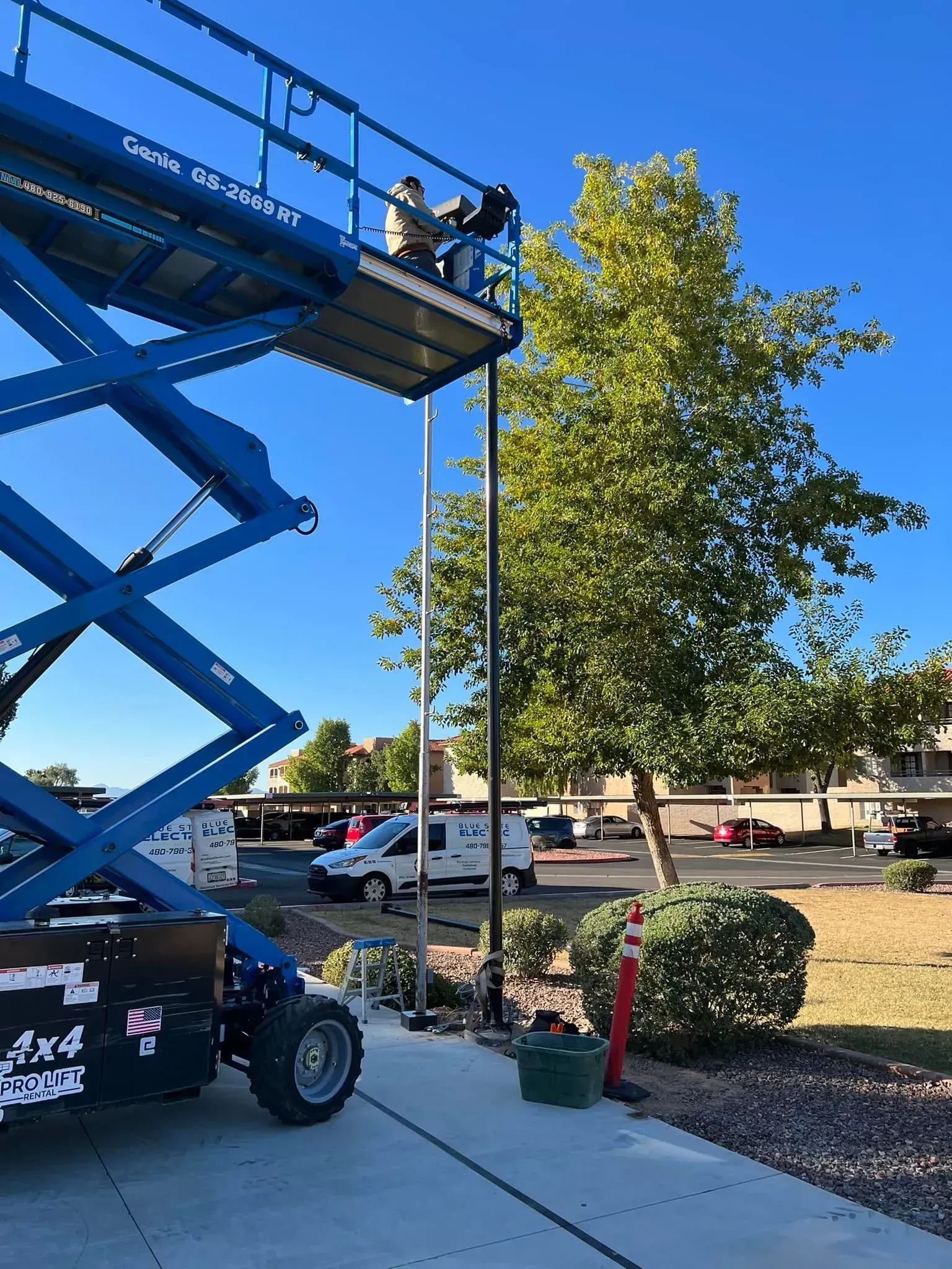 Blue scissor lift beside a tree in a parking lot, with two workers on the platform.