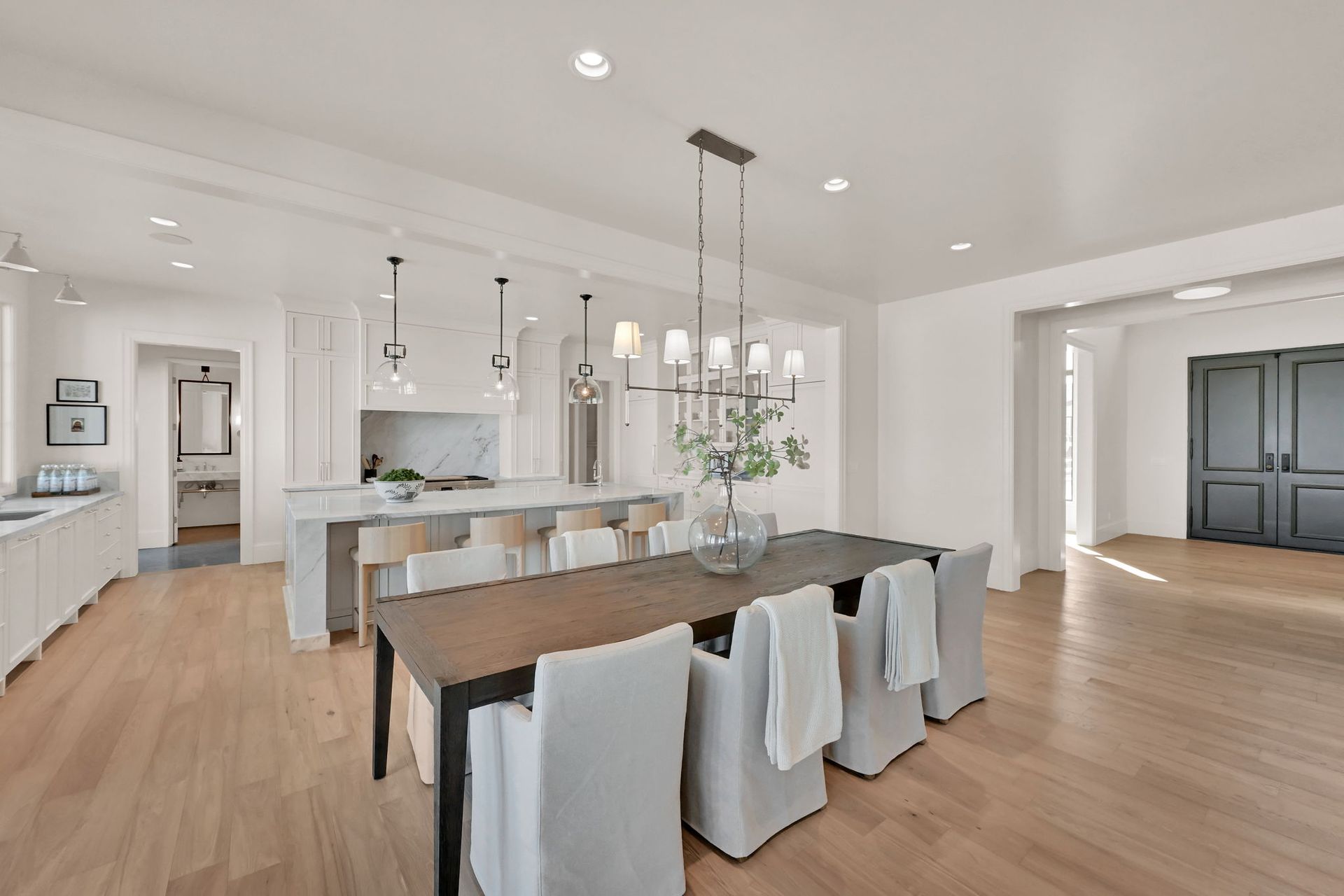 A dining room with a long table and chairs and a kitchen in the background.