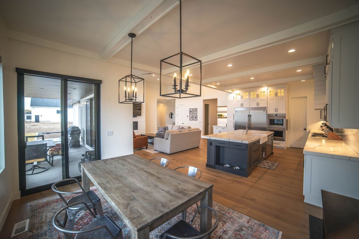 A dining room with a wooden table and chairs and a kitchen in the background.