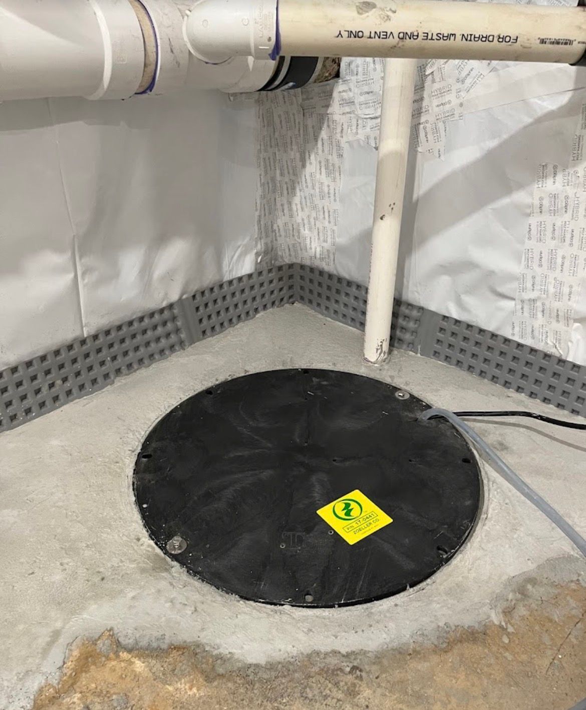 A black sump pump cover in a concrete basement floor, with pipes and wall lining visible.