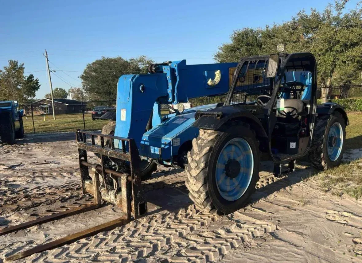 Blue telehandler on a dirt surface with forks extended, under a blue sky.