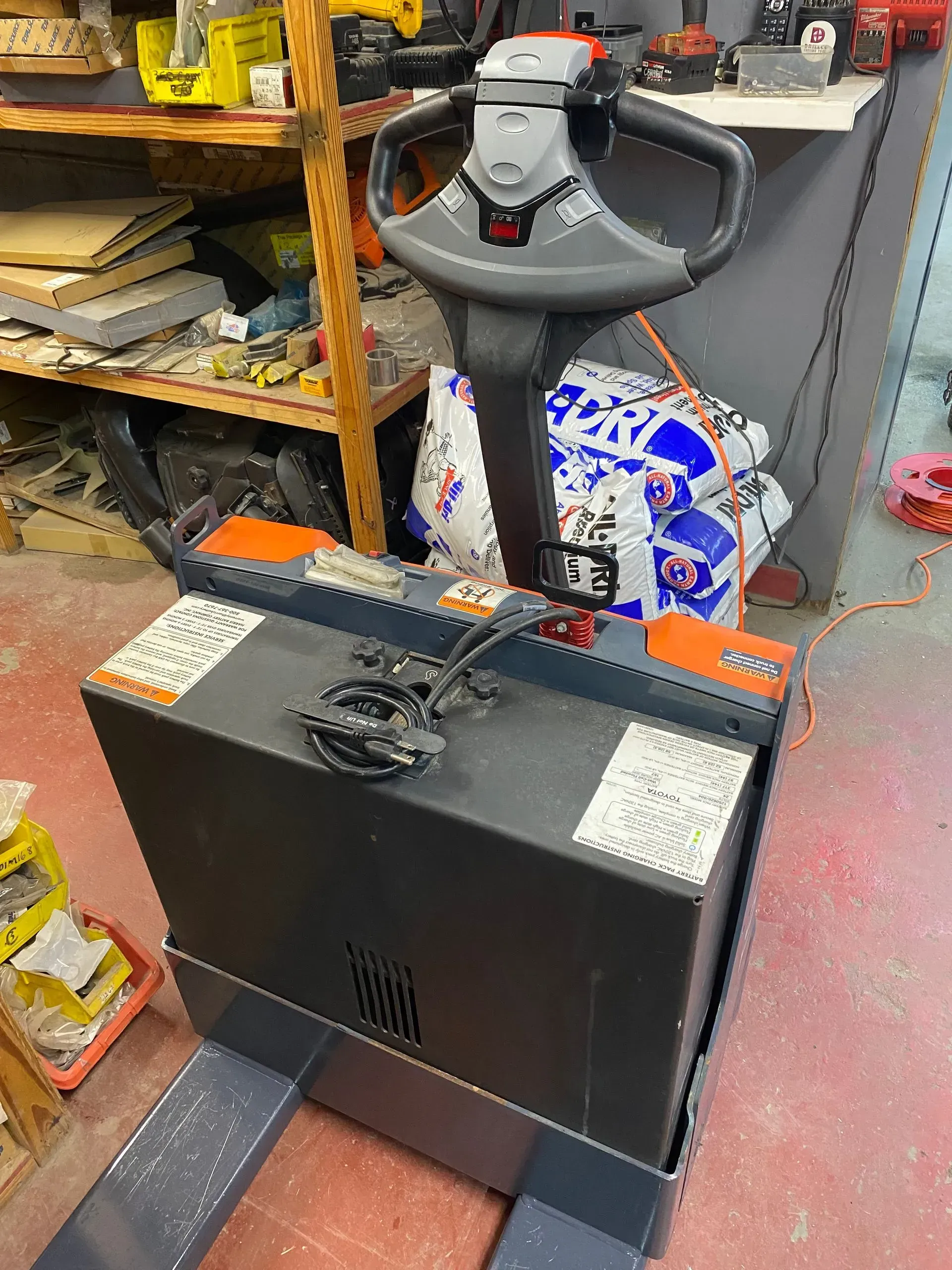 Electric pallet jack with a gray control handle and a black battery case in a workshop.