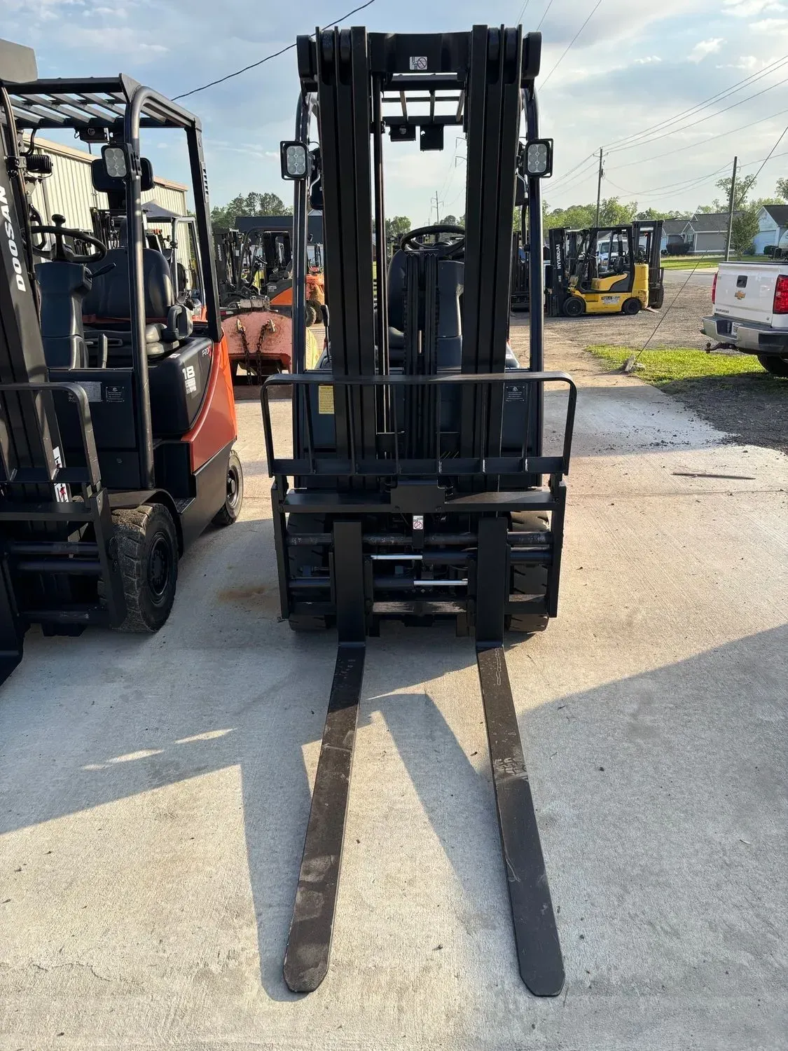 Black forklift with long forks on a paved lot, other forklifts and buildings in the background.