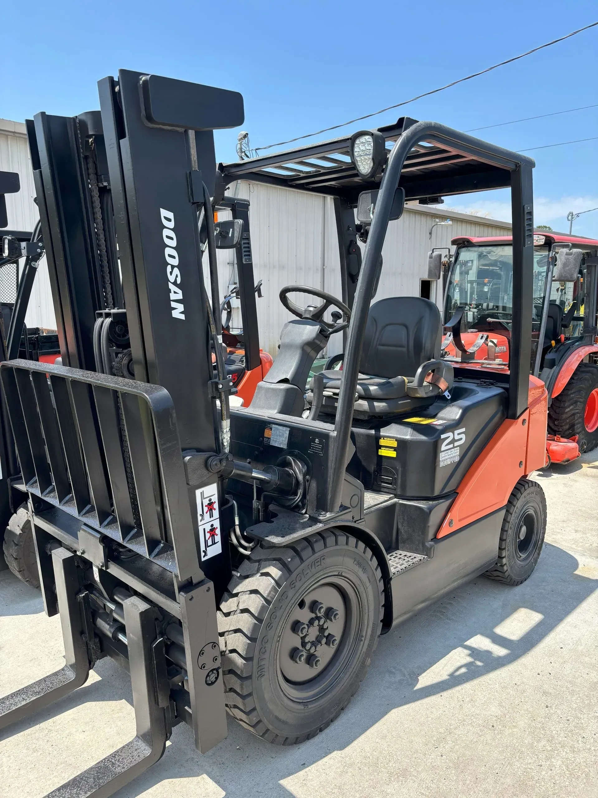 A Doosan forklift, orange and black, parked outside, with a tractor visible in the background.