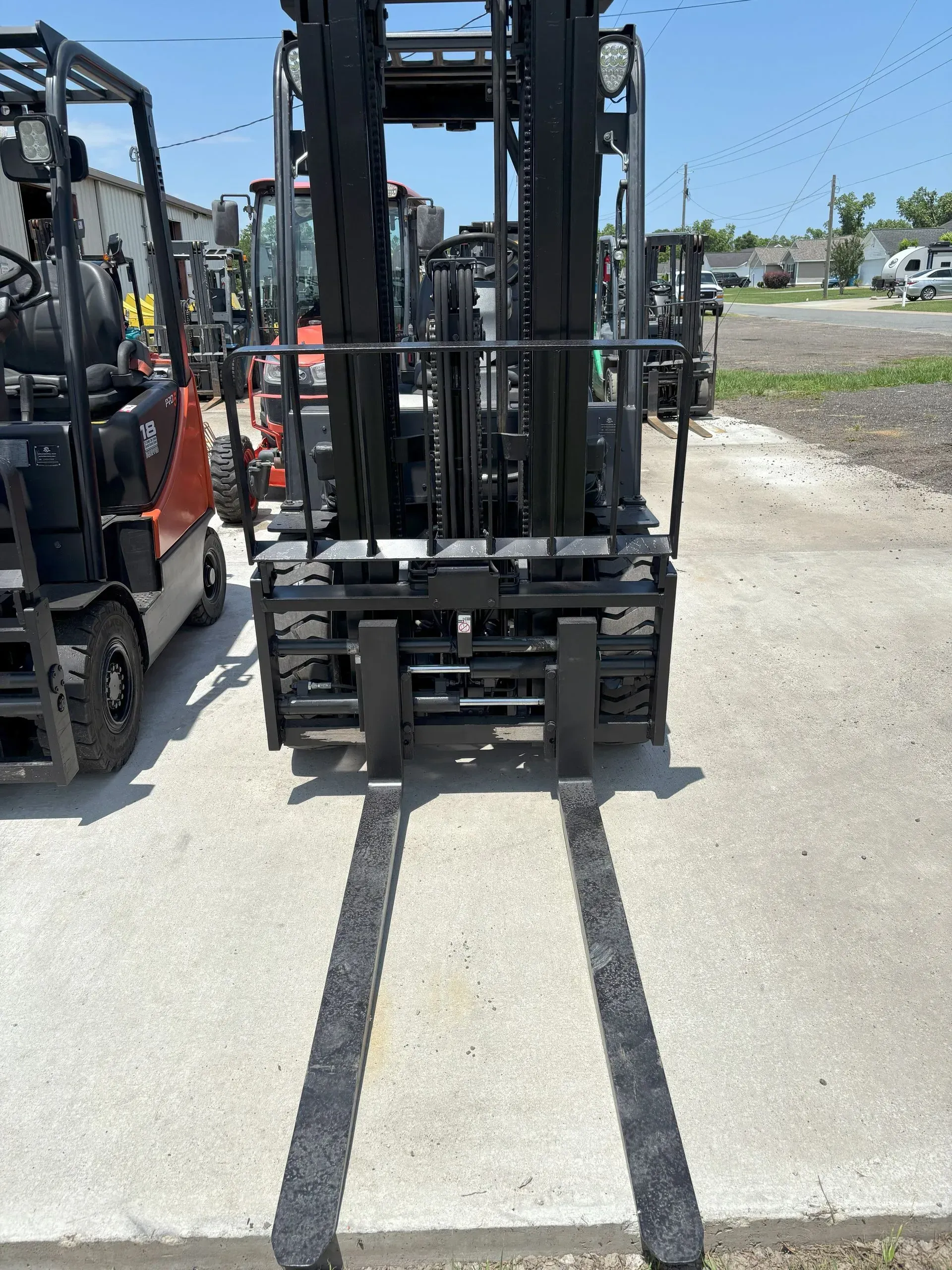 Forklift on a concrete surface with two visible tines, other machinery in the background on a sunny day.