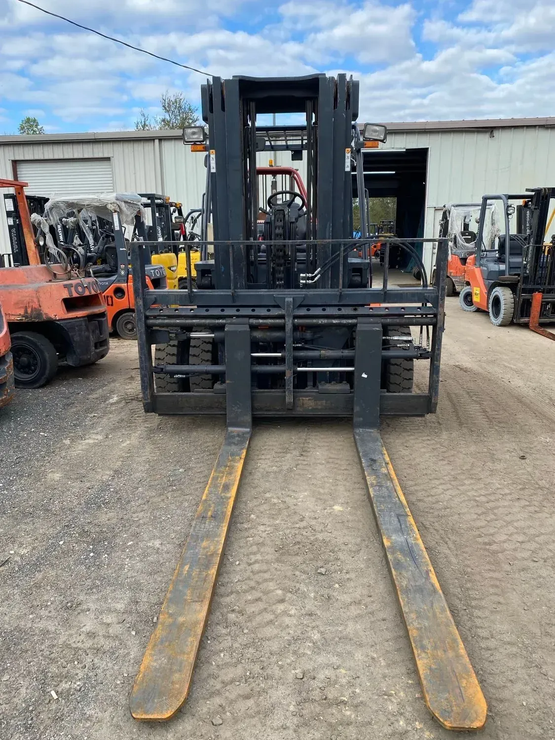 A forklift with long, yellow-tipped forks is parked outside a warehouse; other forklifts visible in the background.