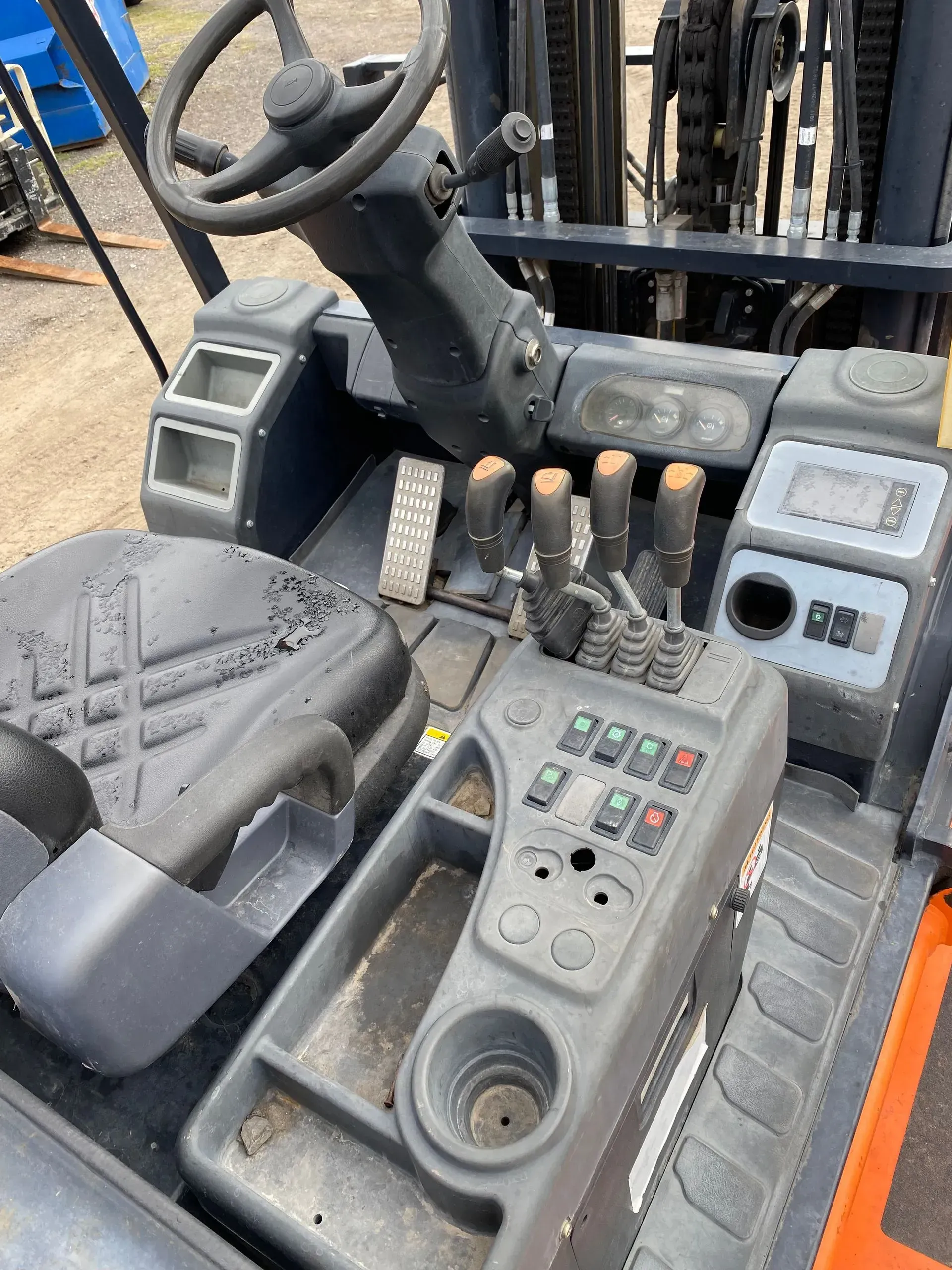 Interior view of a forklift cab with steering wheel, levers, pedals, and control panel. Black and gray.