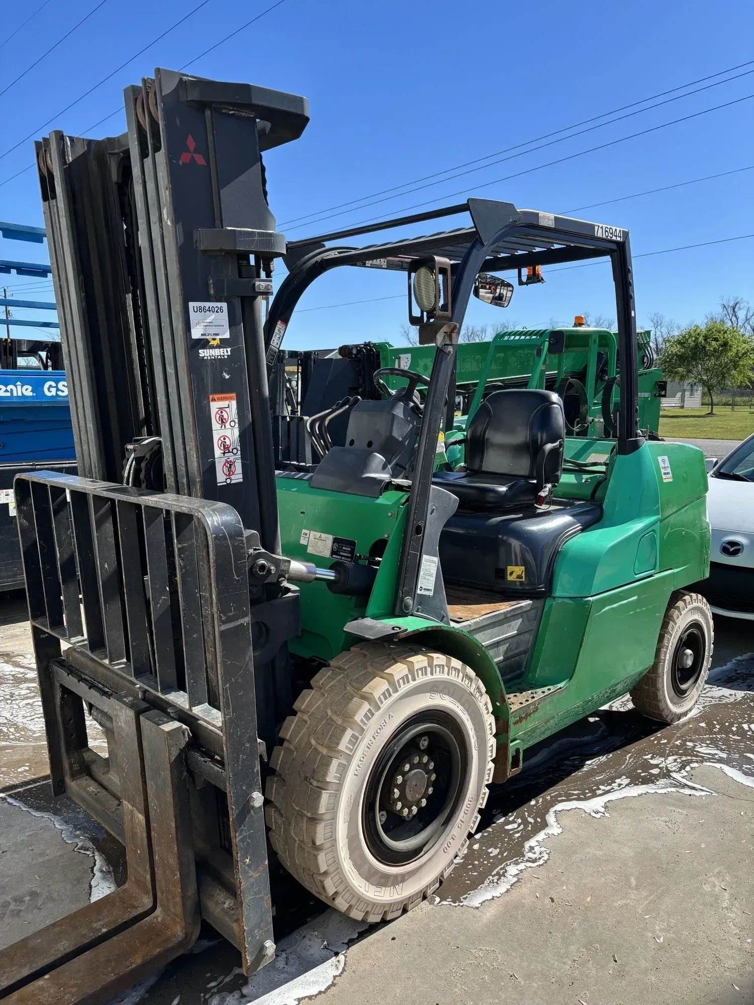 Green Mitsubishi forklift on a paved lot under a blue sky.