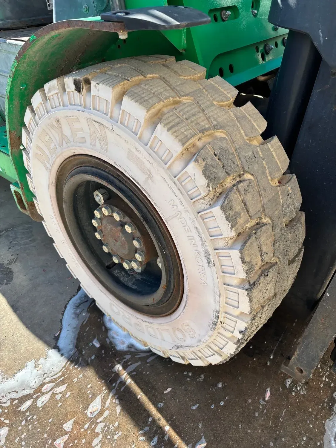 Forklift tire, off-white, with dark metal wheel. Green forklift body in the background, set outside.