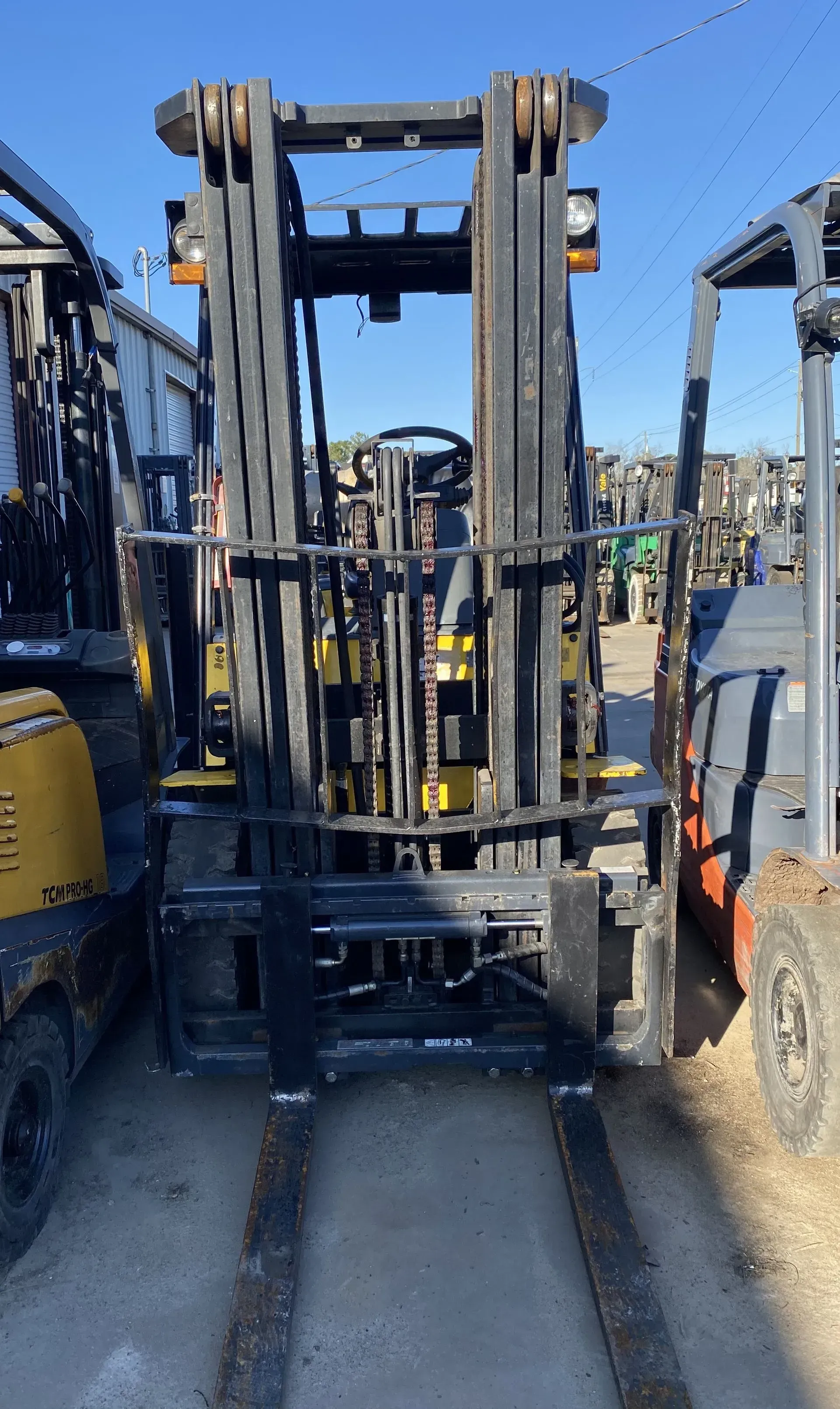 Black and yellow forklift with forks extended, parked between other forklifts in a lot.