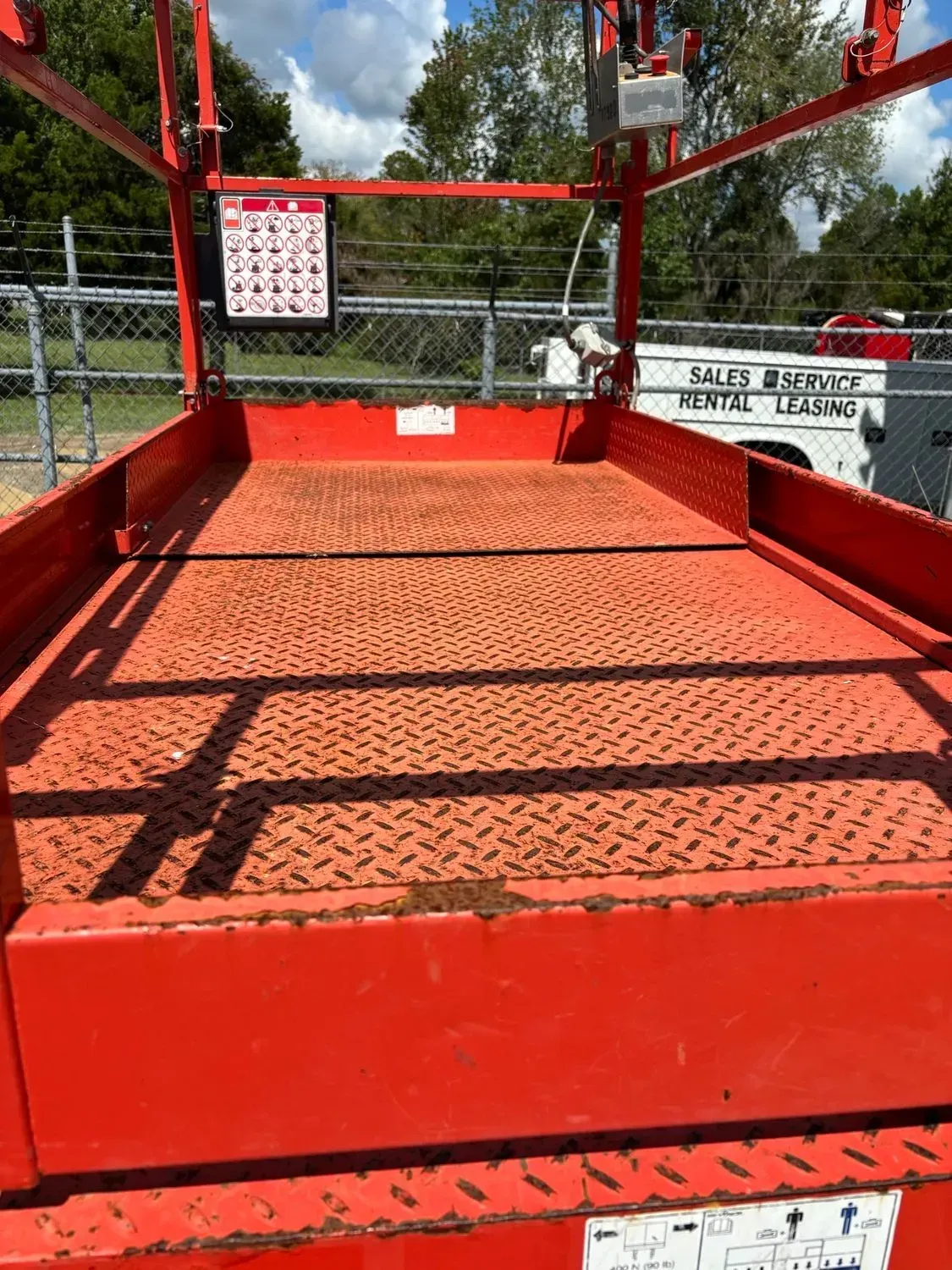 Red scissor lift platform, empty. Safety instructions visible. Outdoors, bright day.
