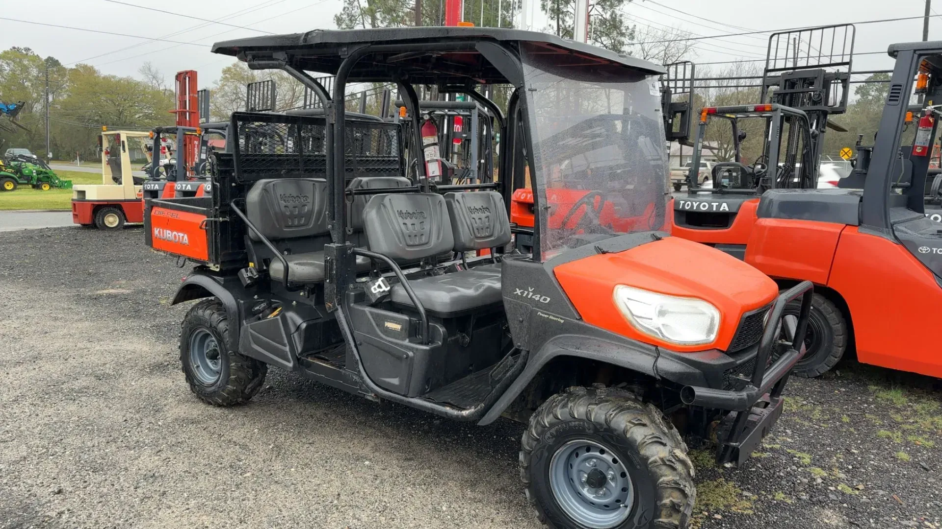 Orange and black Kubota utility vehicle with a canopy, parked outside, with forklifts in the background.