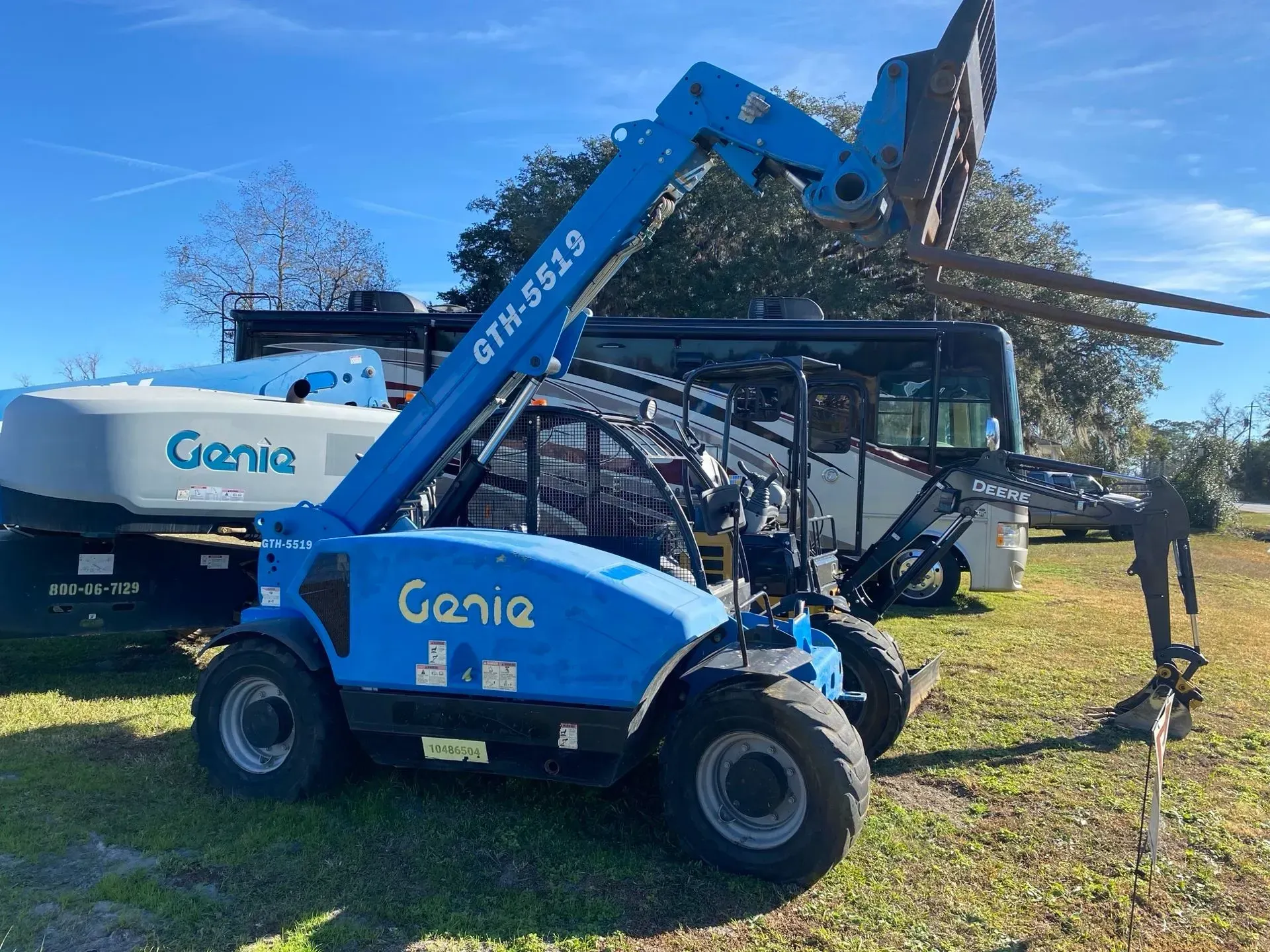 Blue Genie telehandler with forks extended, parked on grass near a vehicle. Sunny day.