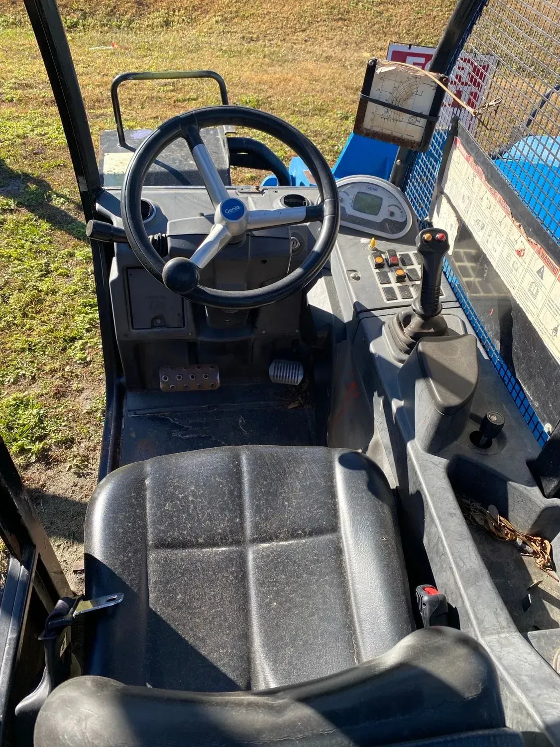 Interior of a utility vehicle: black steering wheel, seat, and controls, blue accents. Outdoor setting.