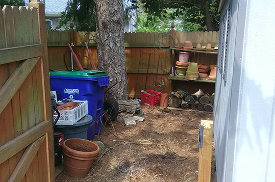 A blue trash can is sitting in a backyard next to a wooden fence.
