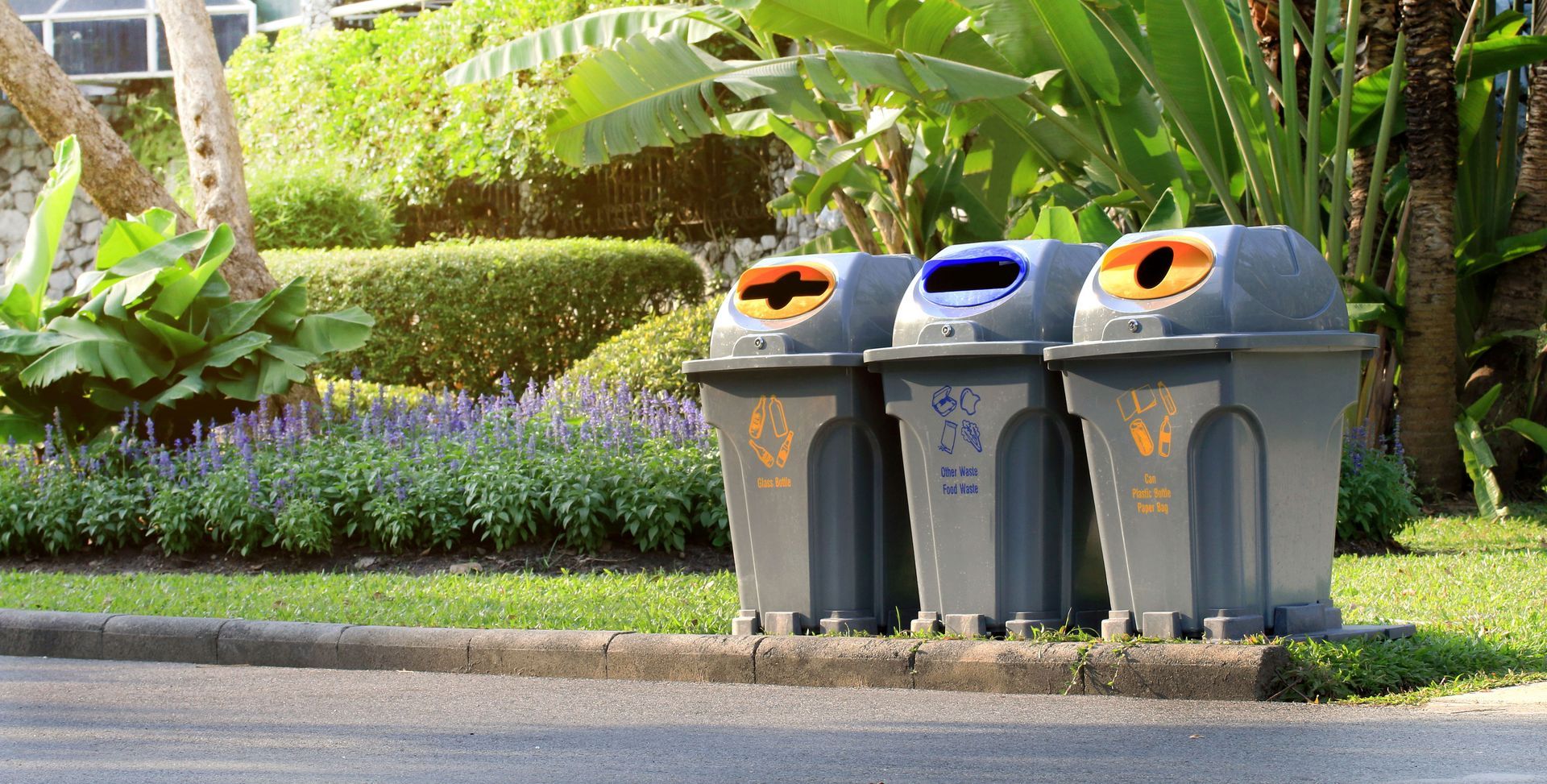 Three recycling bins with orange, blue, and orange lids sit on a curb next to a garden.