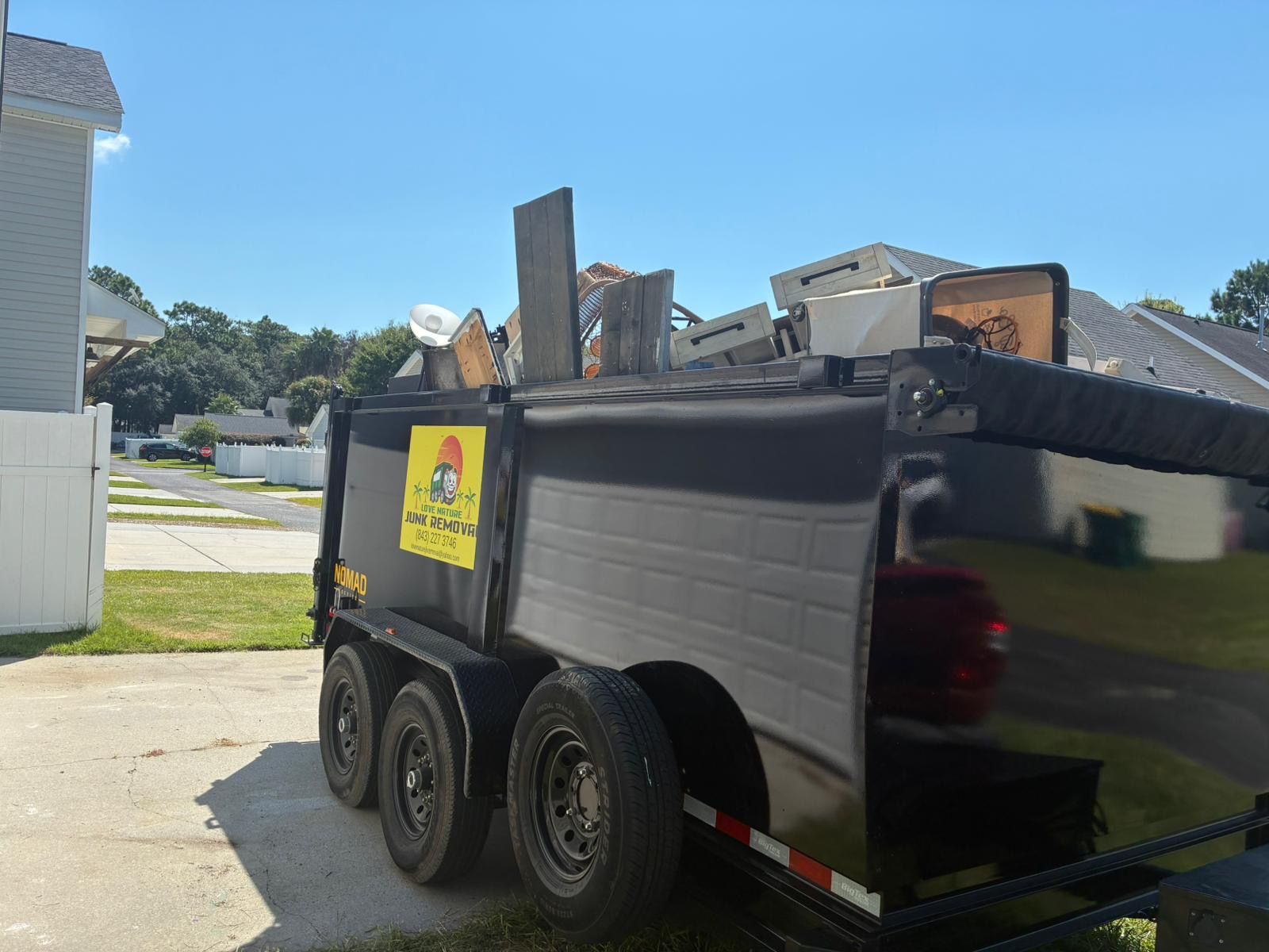 Black trailer filled with debris parked on a driveway in front of a house on a sunny day.