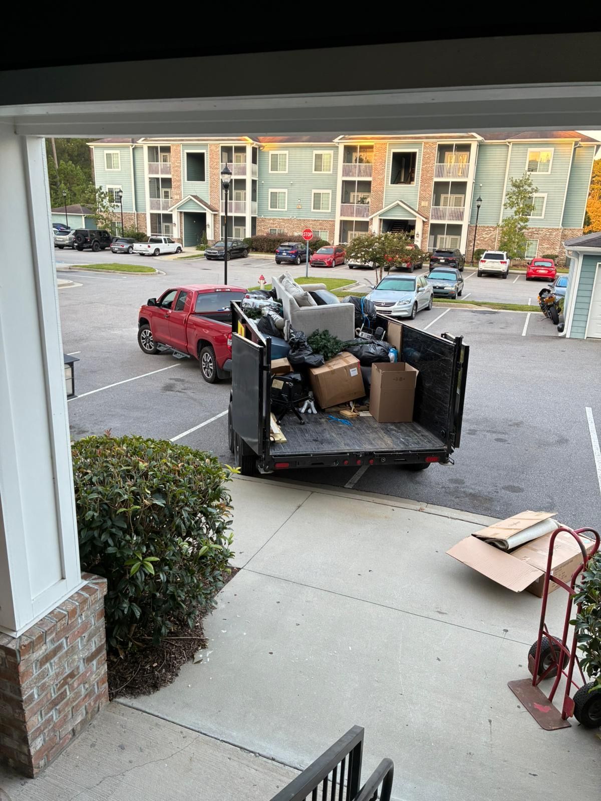 A truck filled with junk sits in front of an apartment complex. Cardboard boxes and furniture fill the truck bed.