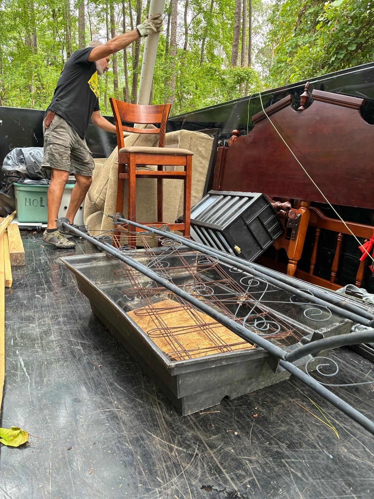 Man loading furniture into a truck: chair, bed frame, box, forest background.