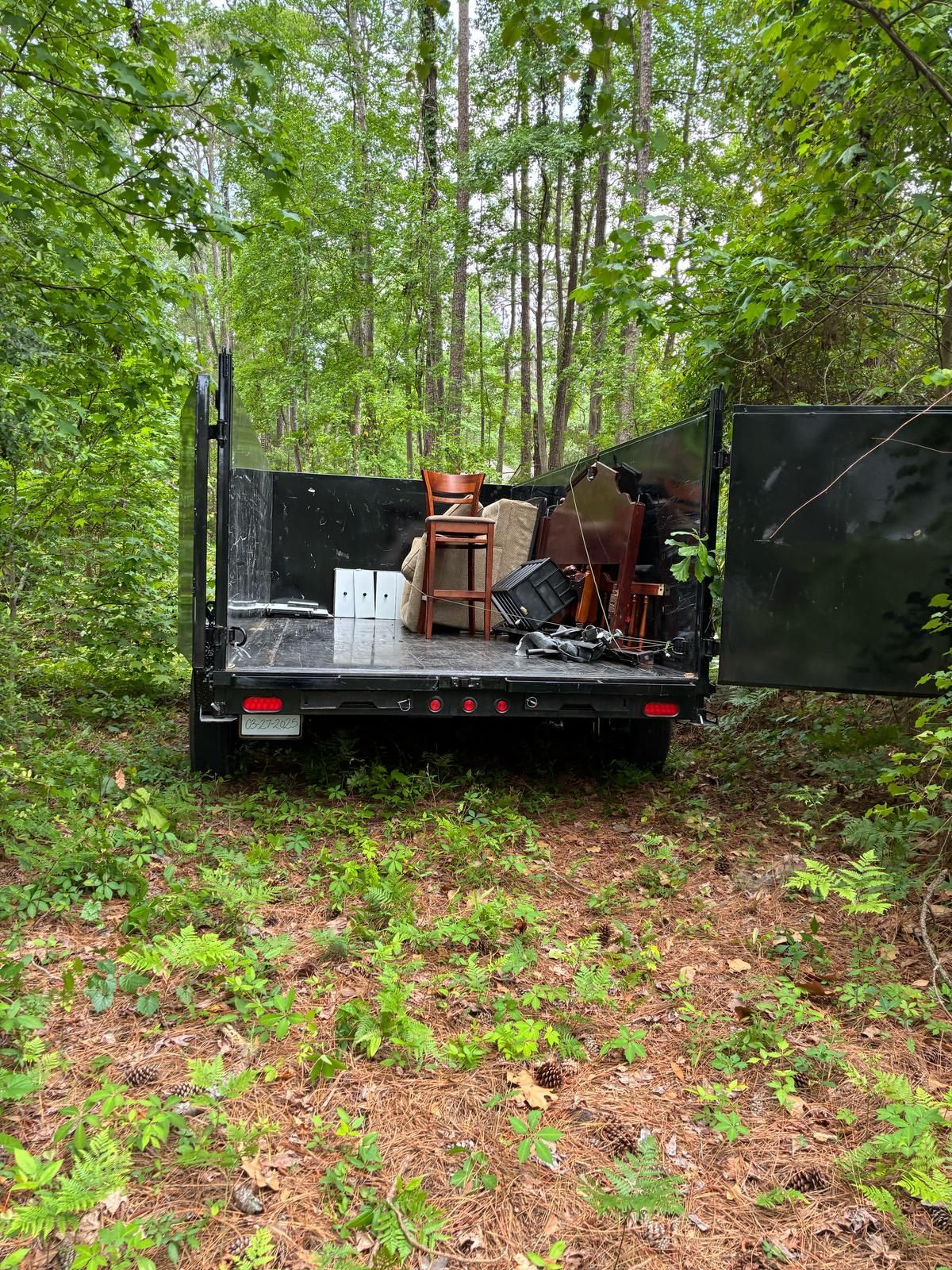 Black truck bed filled with furniture, parked in a wooded area with tall trees and green foliage.