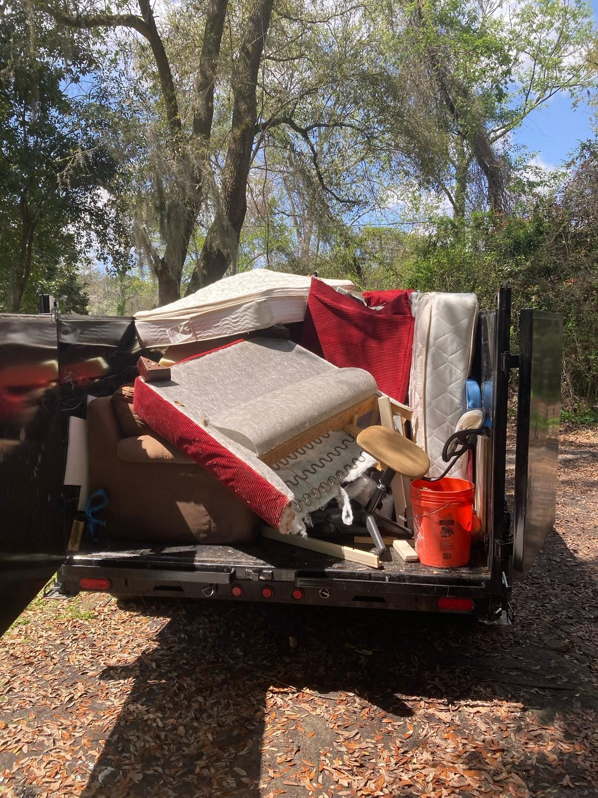 Trailer filled with furniture and debris in a wooded area. Orange bucket visible.