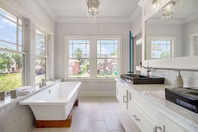 A bright, modern bathroom featuring a white clawfoot bathtub under a large window and a long white double vanity.