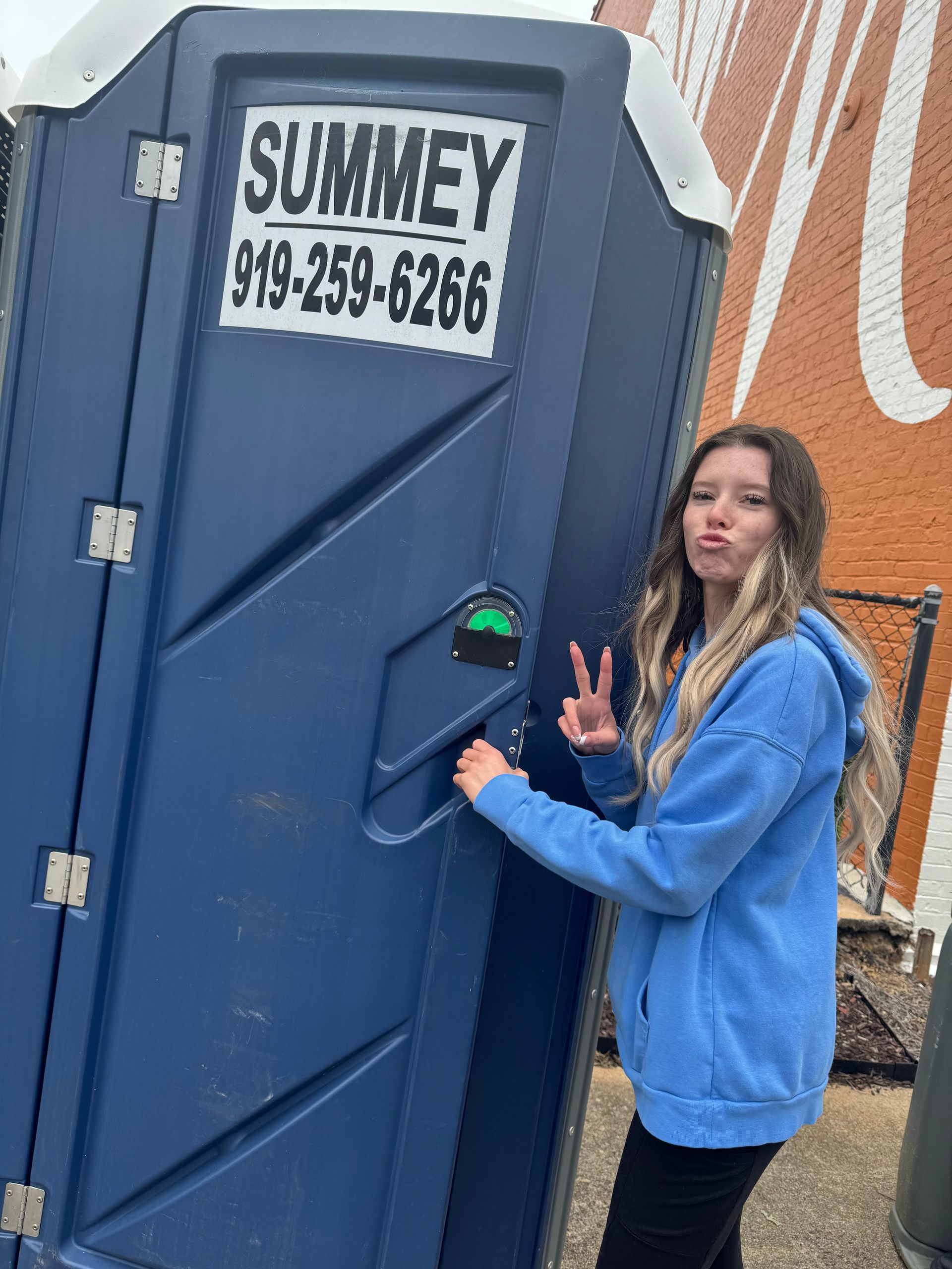 A woman in a blue hoodie is standing next to a blue portable toilet.