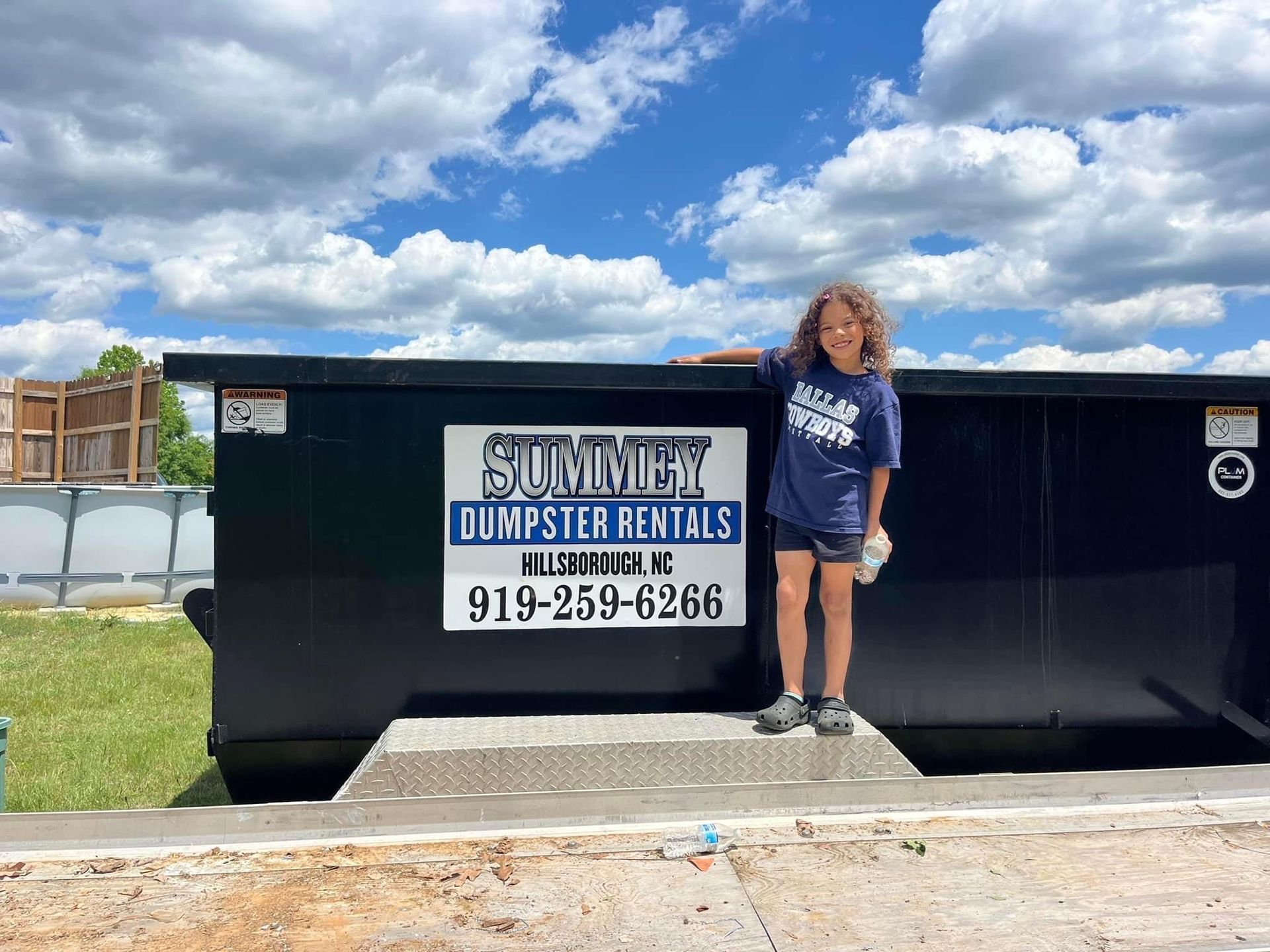 A young girl is standing in front of a dumpster.