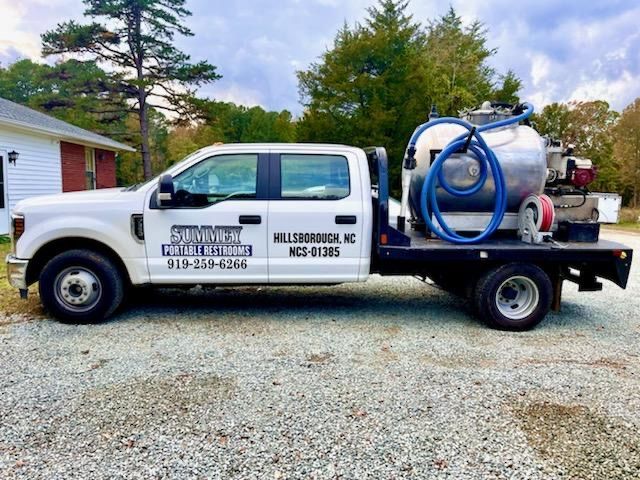 A white truck with a vacuum tank attached to the back is parked in a gravel lot.