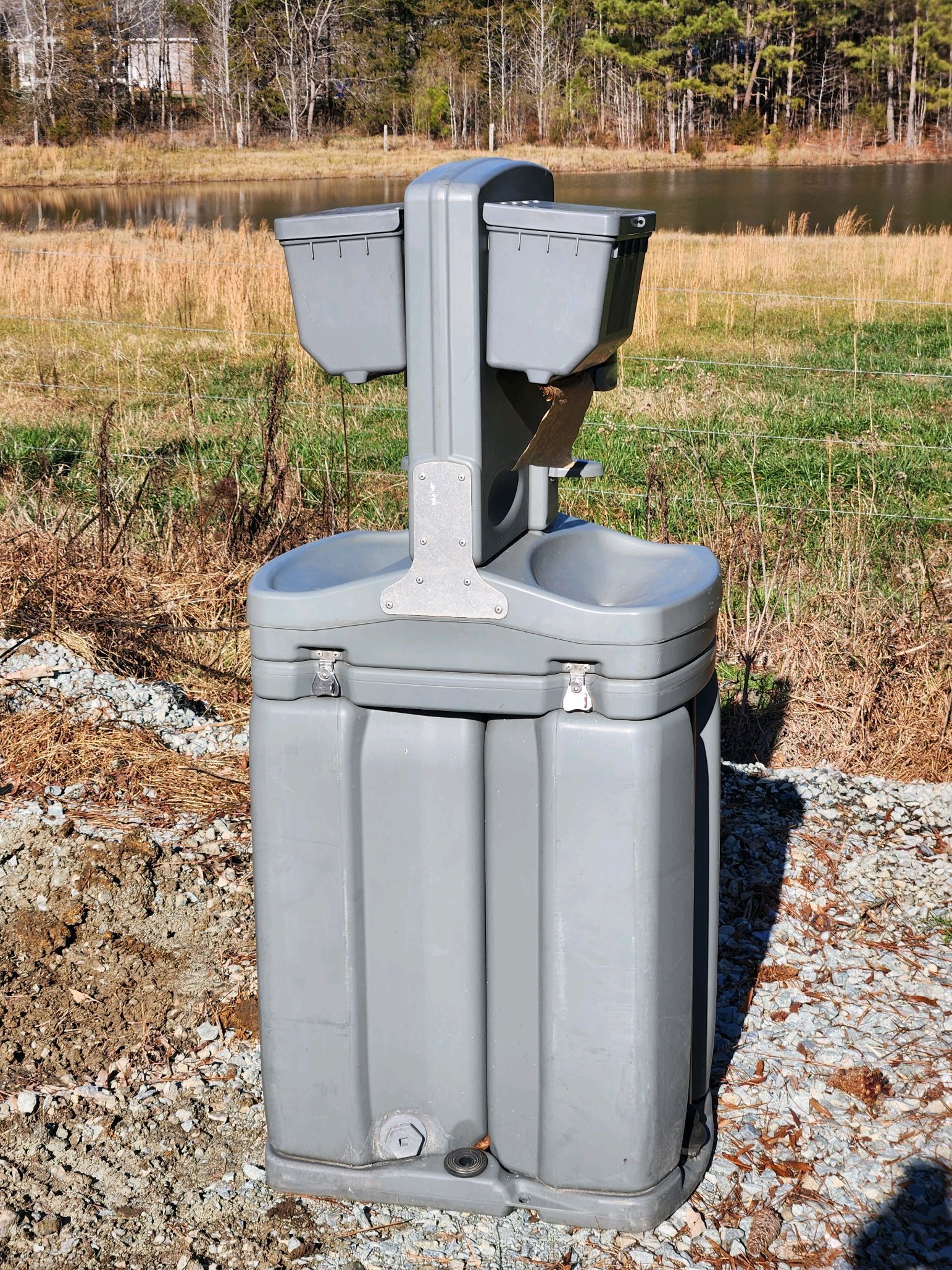 A gray trash can is sitting on a gravel road next to a body of water.