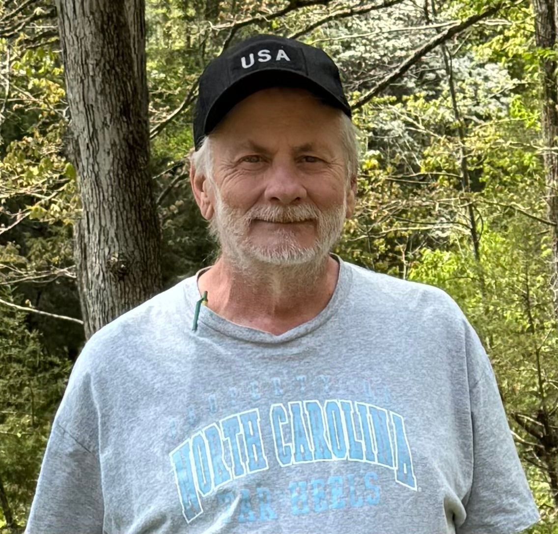 A man wearing a hat and a gray shirt is standing in the woods.