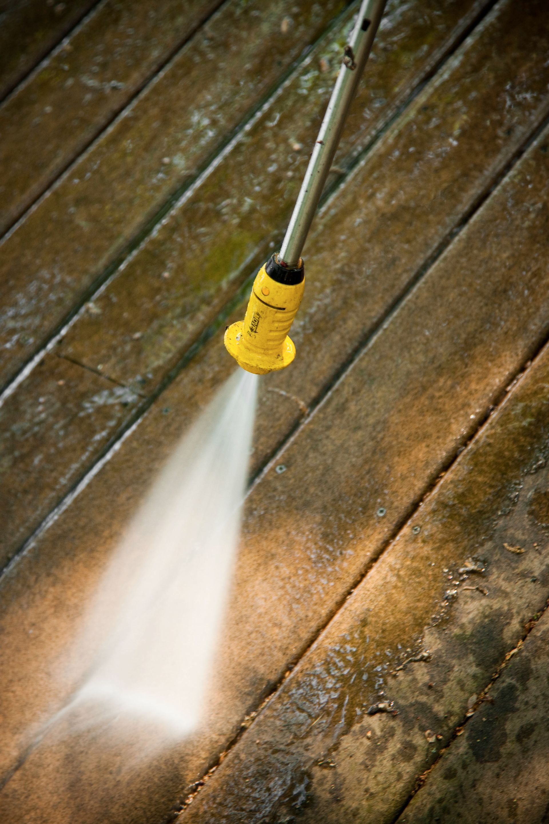 Pressure washer spraying a wooden deck, removing grime. Yellow nozzle, angled downwards.