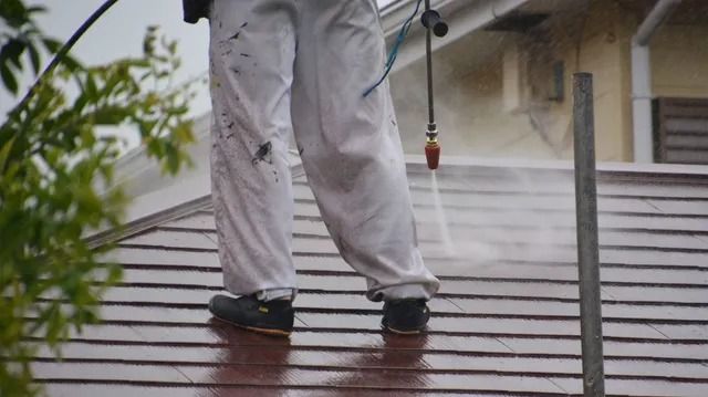 Person in white coveralls power washing a brown shingle roof, spraying water.