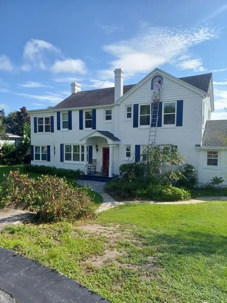 White two-story house with blue shutters; person on a ladder painting; green lawn and blue sky.