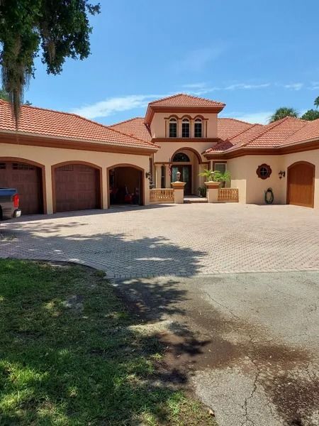 Tan stucco house with terracotta roof, three-car garage, and brick driveway on a sunny day.