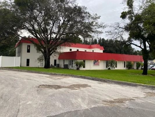 White building with red roof, American flag, on a grassy area. Paved road in foreground.