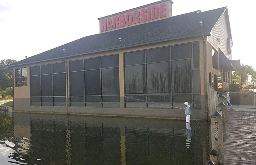Harborside restaurant on water. Beige building with dark tinted windows, dark roof. Sign on top.