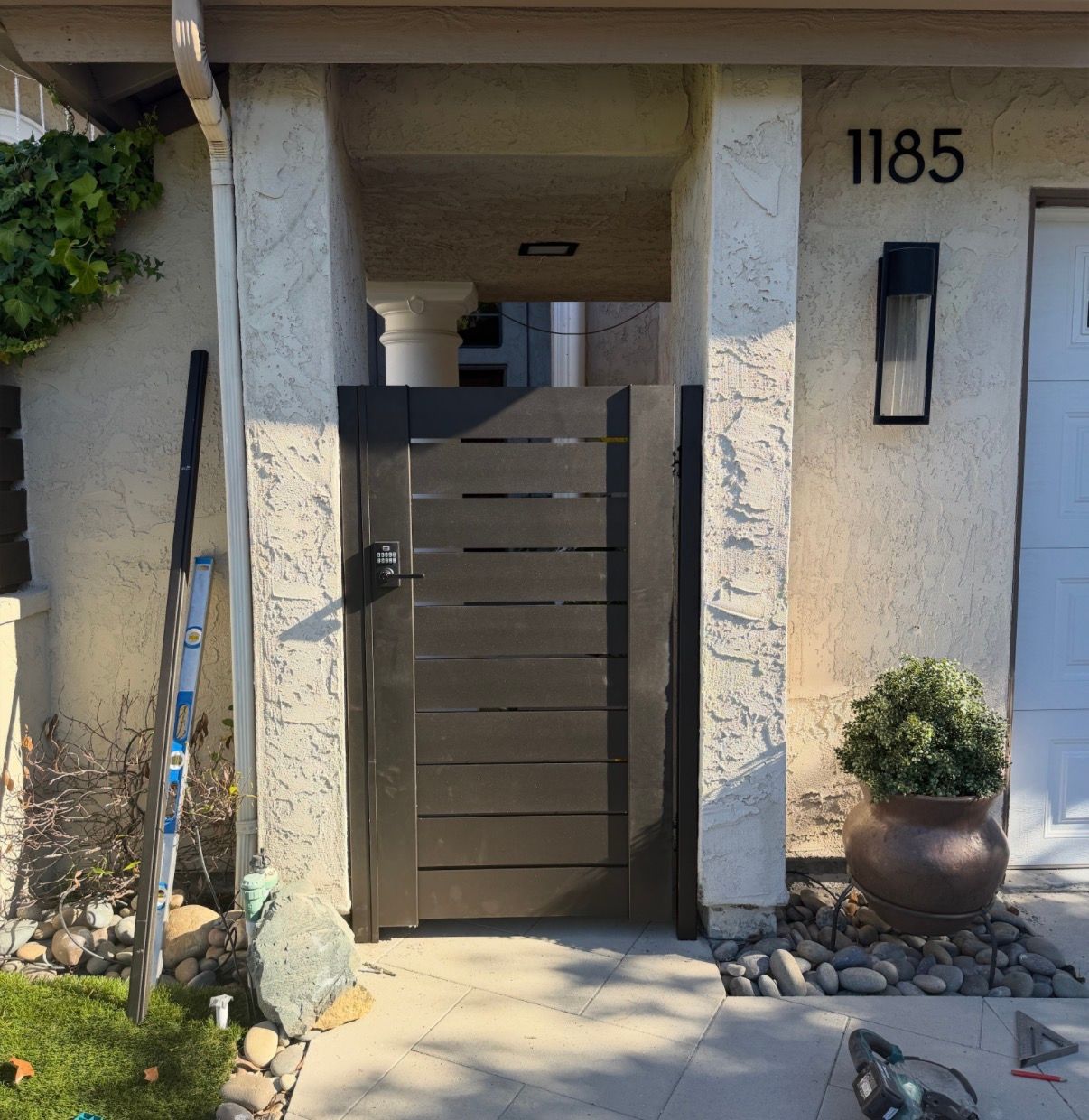 Modern house entrance with a dark gate, stone columns, potted plant, and house number 1185