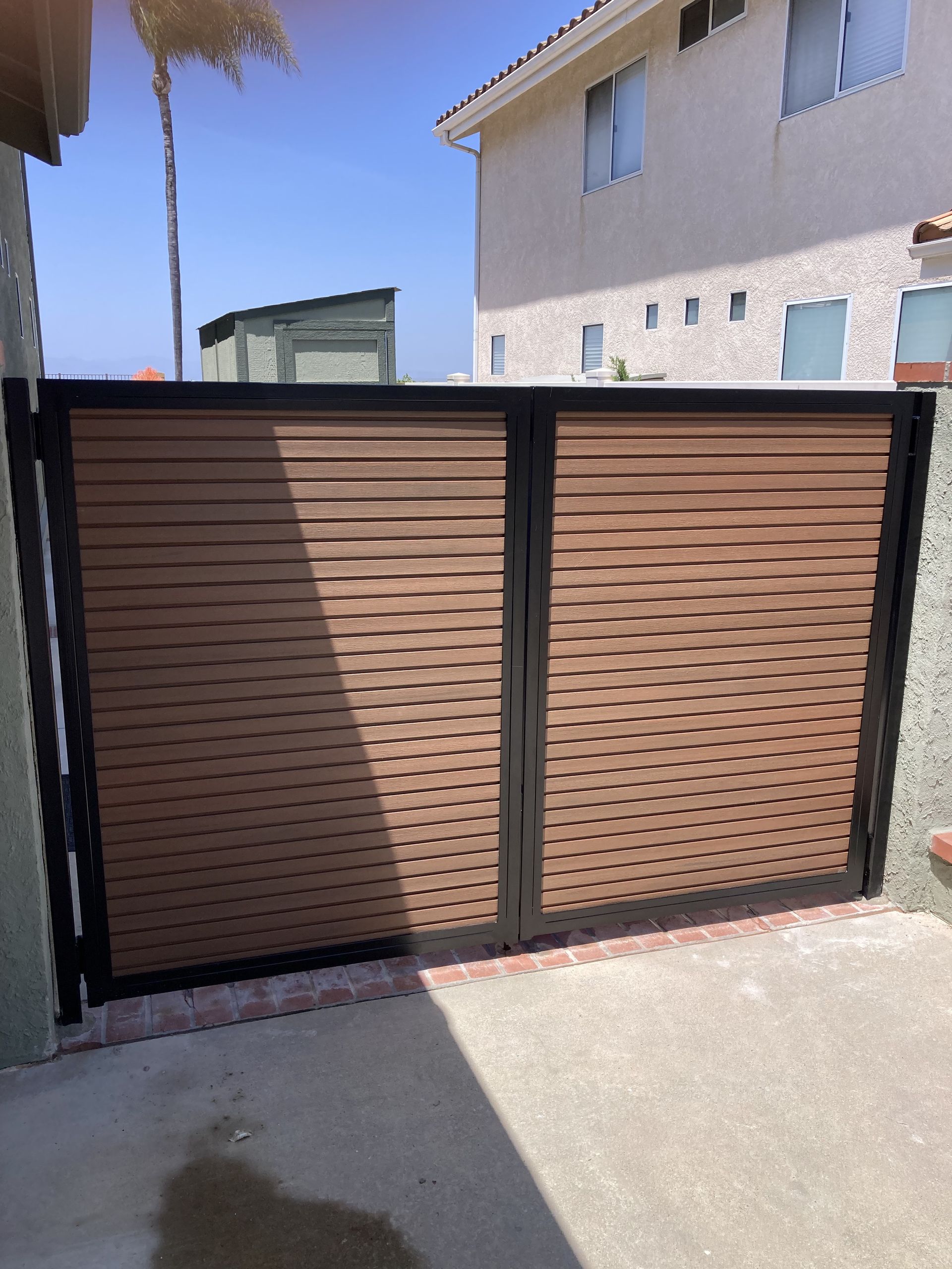 Brown metal driveway gate with horizontal slats beside a beige house in bright sunlight