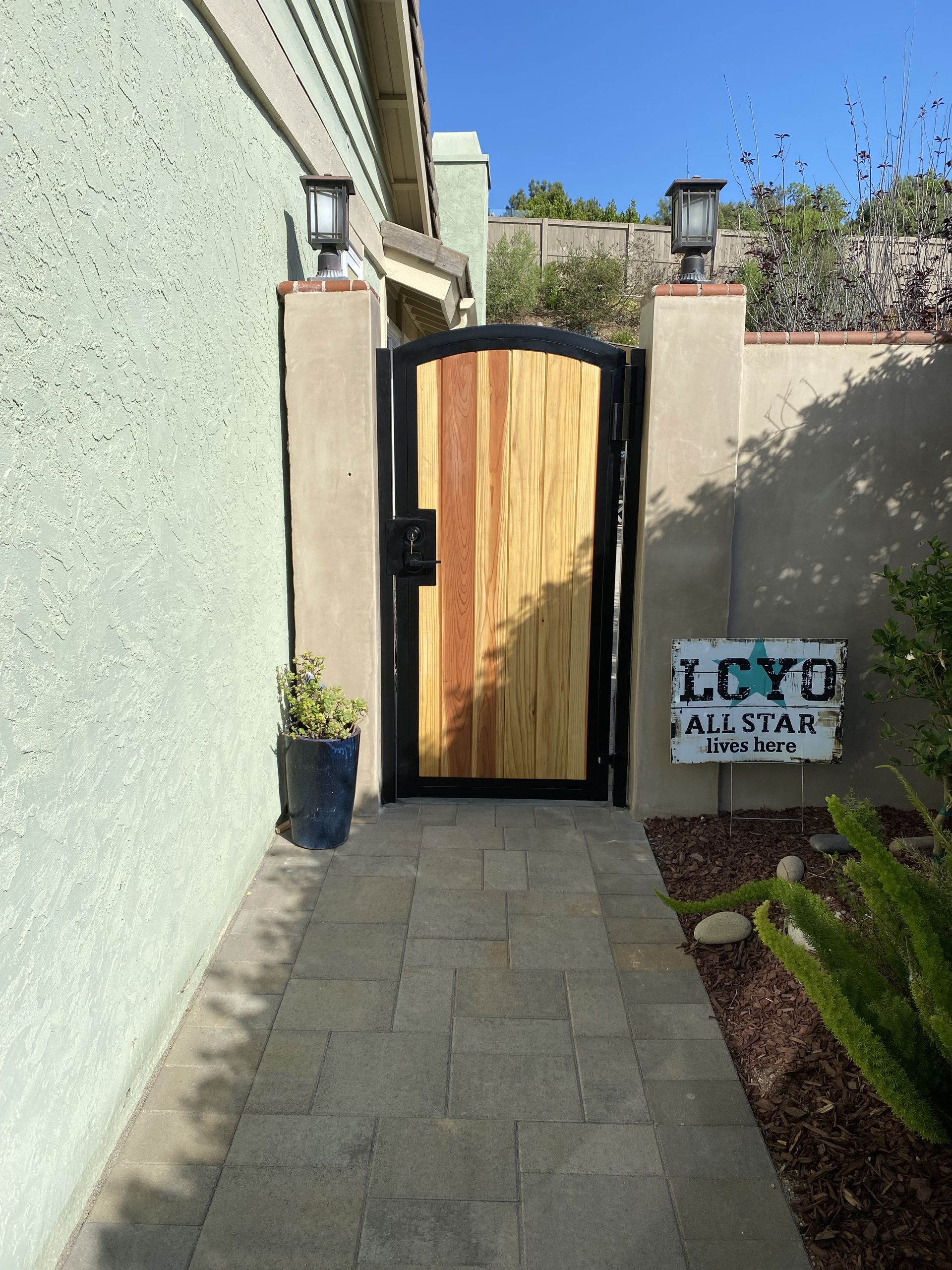 Narrow side yard with stone path leading to a black gate and wooden door, beside a wall sign reading “LUXYQ ALL STAR.”