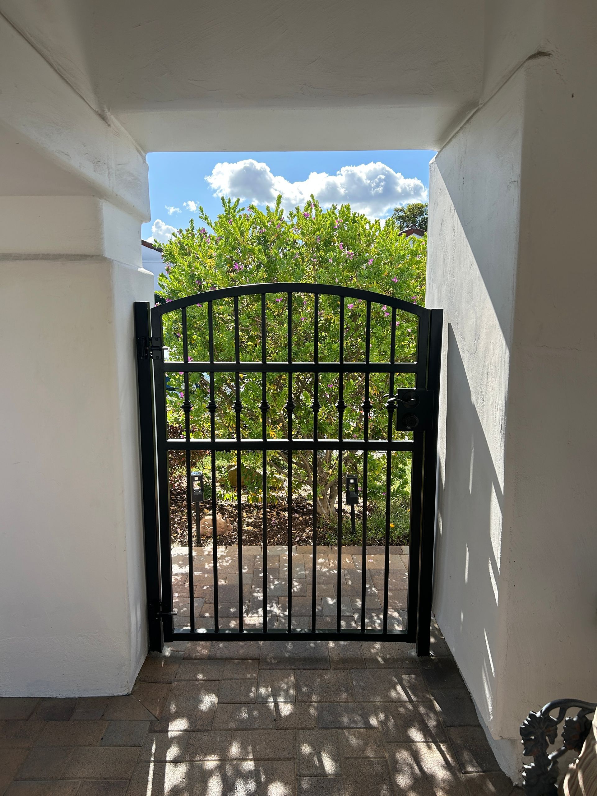 Black metal gate at a white-walled entrance, opening to a sunny path with green trees beyond.
